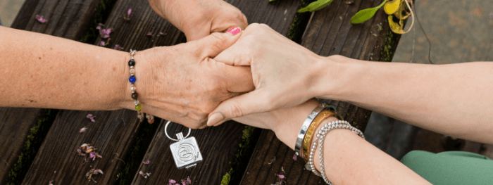 Two people holding hands, one wearing a Sharing Solace locket and support bracelets, symbolizing comfort and connection.