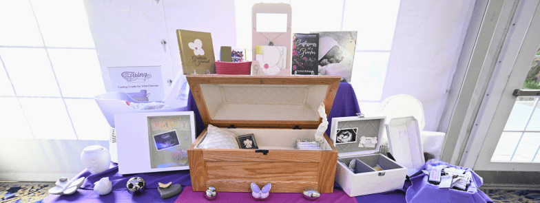 A display table of bereavement keepsakes, including an open wooden infant casket, framed baby photo urns, and small remembrance tokens, arranged on purple cloth.